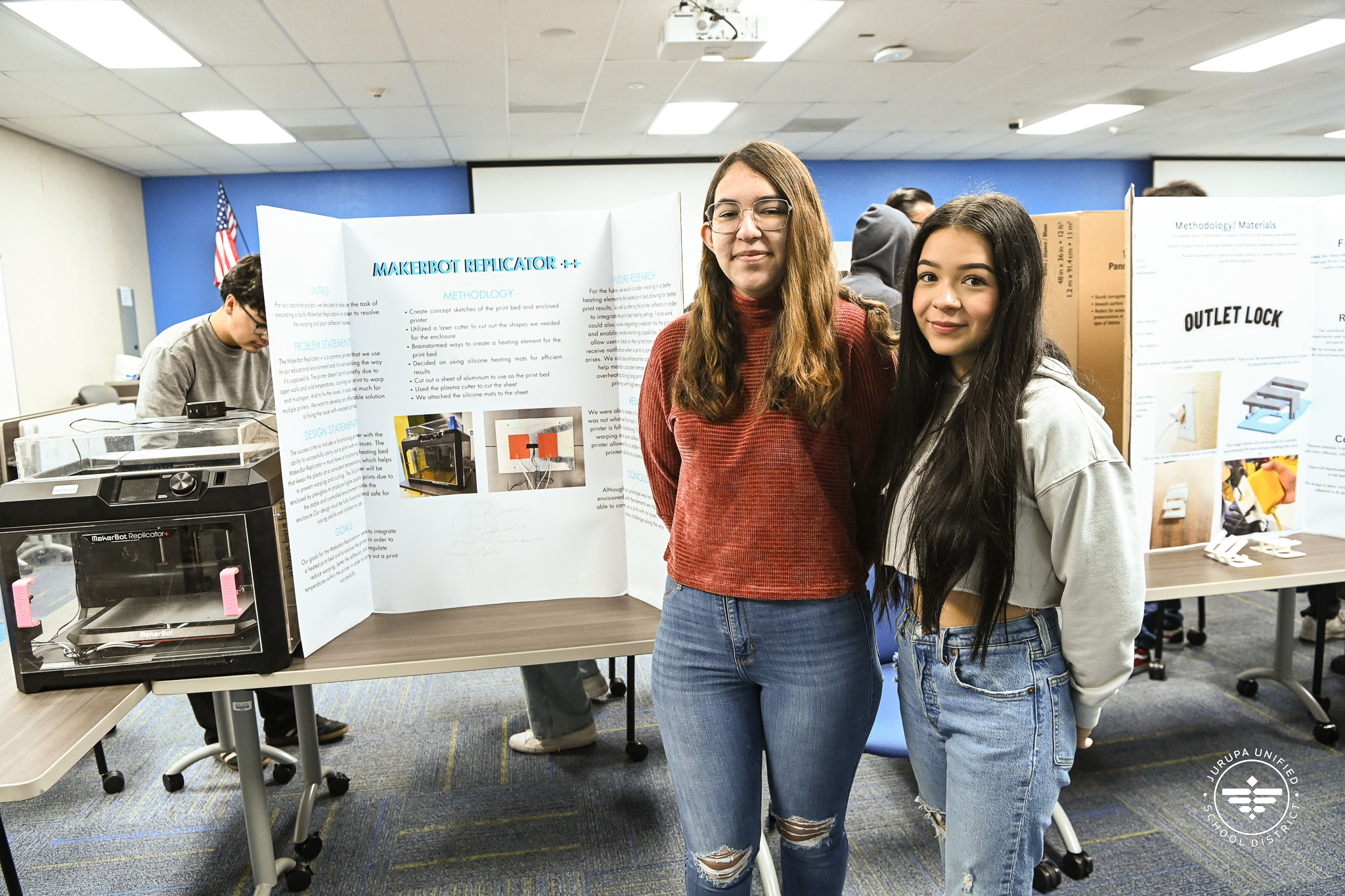 Two students posing in front of their project demonstration and a 3D printer